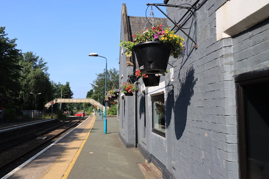 A close-up view of hanging flower baskets attached to a grey brick building at New Barnet station platform. The baskets are filled with vibrant pink, yellow, purple, and white flowers, with green foli