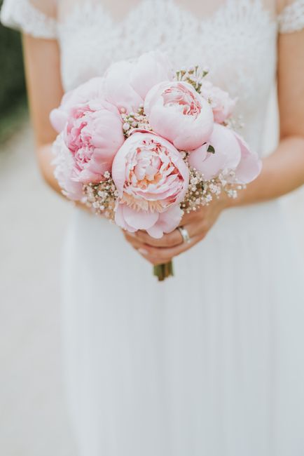 A bride dressed in a white lace gown is holding a bouquet of large, soft pink peonies, accented with small white baby's breath and green foliage. The bouquet features lush, rounded blooms with ruffled