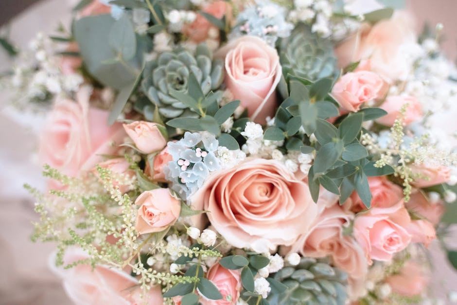 A close-up view of a floral bouquet featuring large, soft pink roses with gently curled petals, complemented by smaller pink spray roses, and accented with light blue succulent foliage. The arrangemen